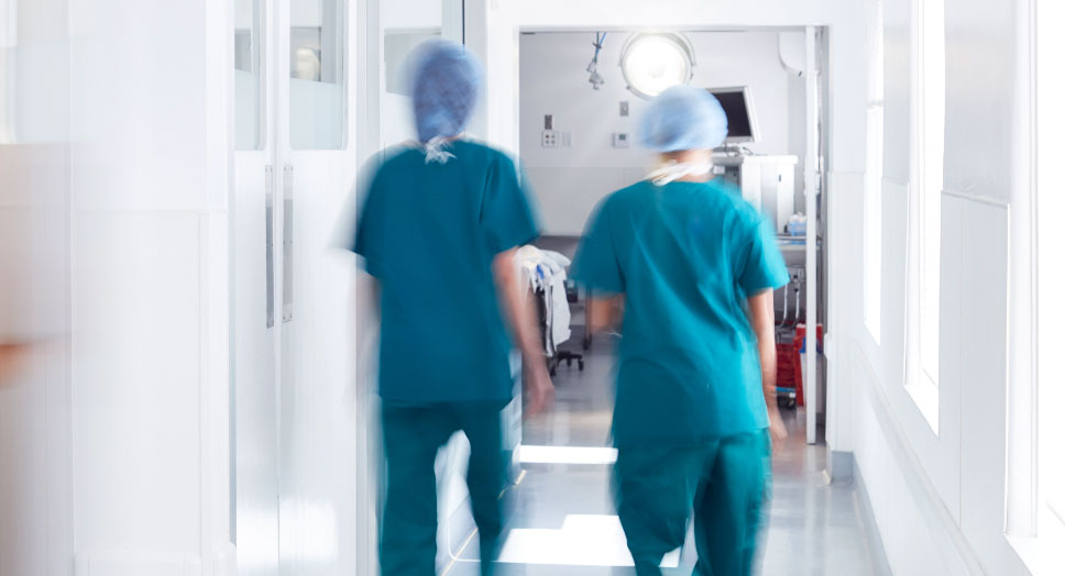Two OR medical workers in blue scrubs and caps walking down a hall.